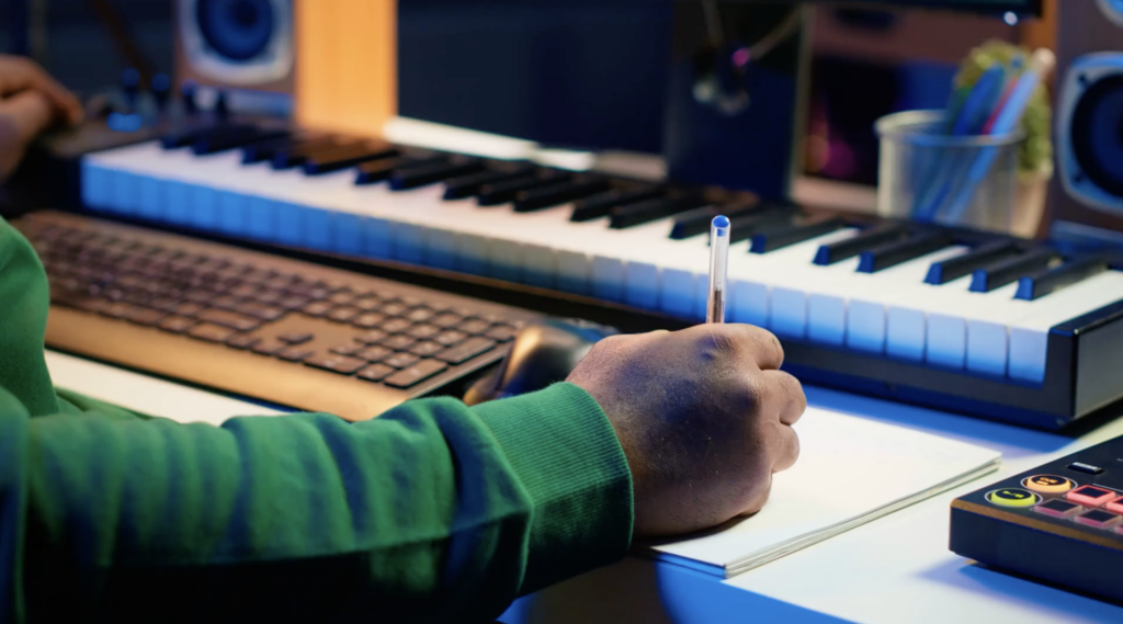 a hand holding a pen in front of a keyboard. SongTown Lyric Writing